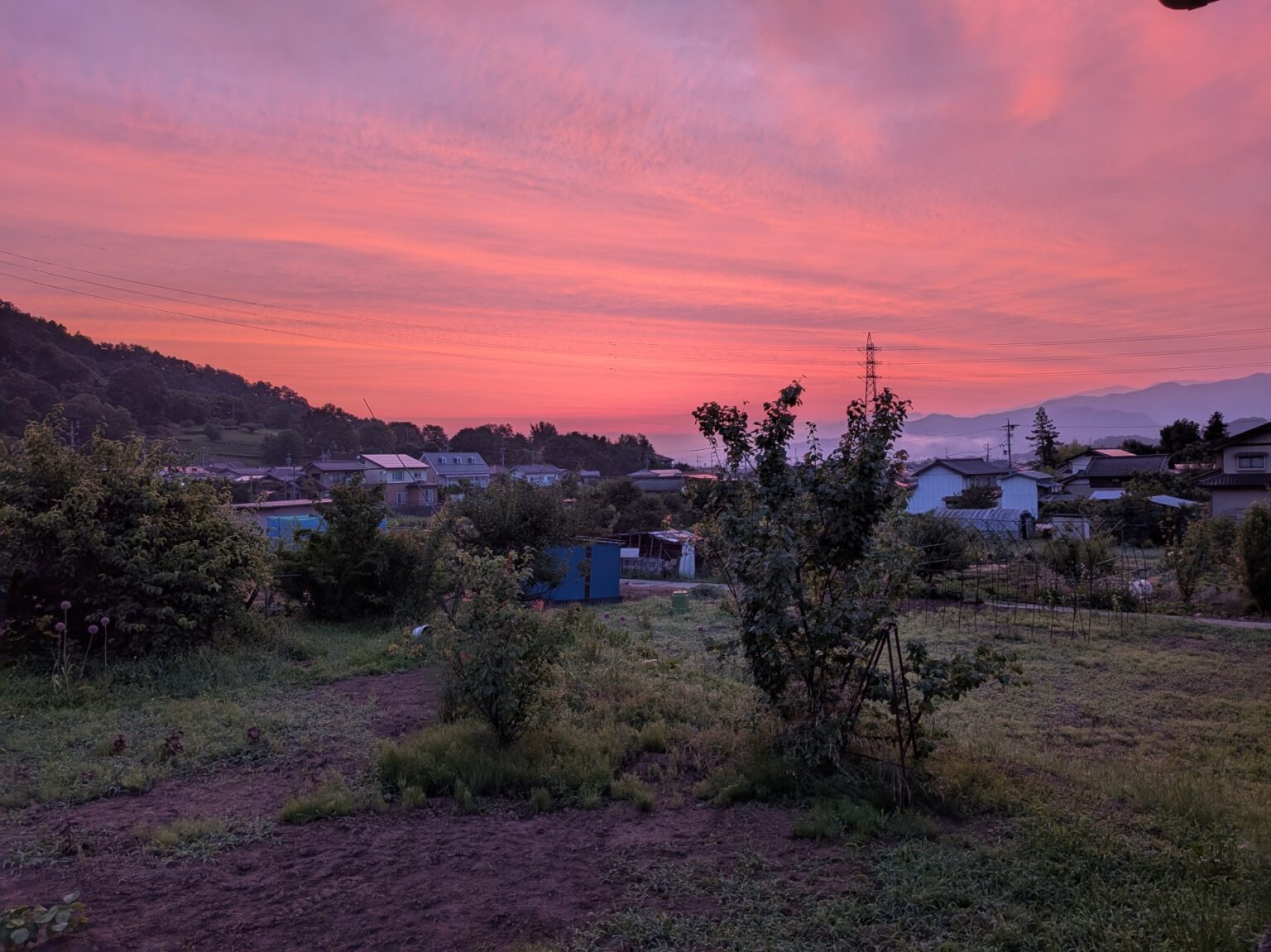 施設周辺の夕焼け風景。田園地帯に建つ複数の建物と山々を望む。